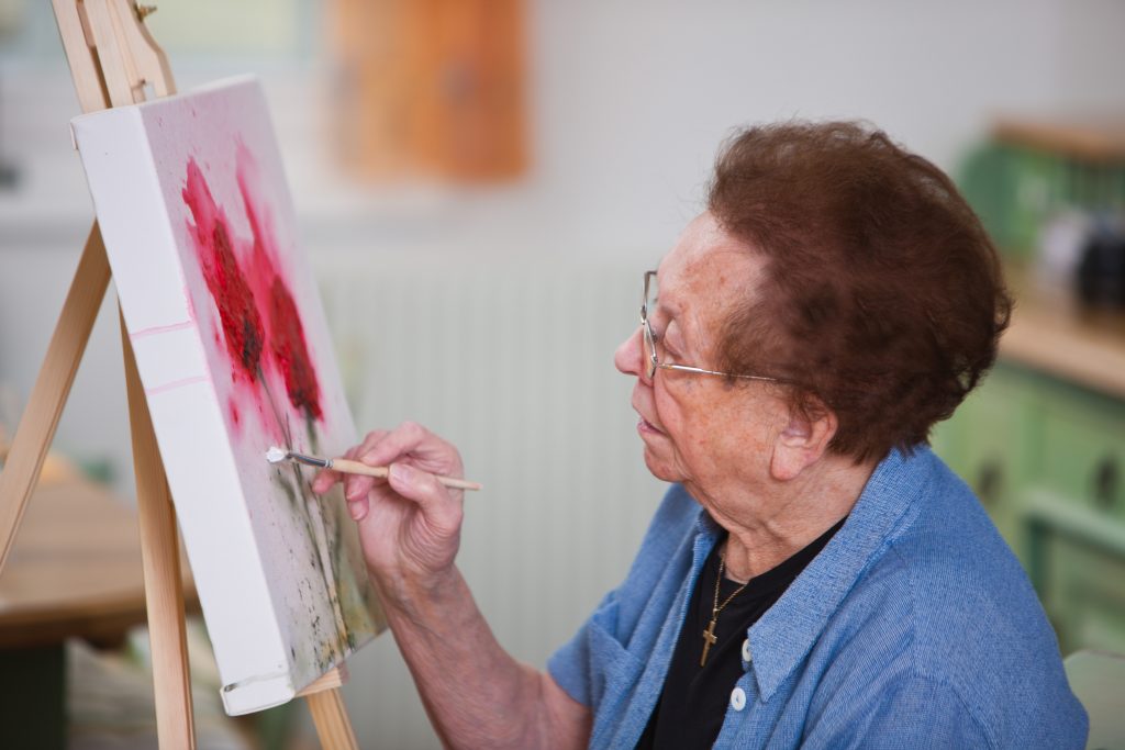 Dôchodkyňa s okuliarmi maľuje červené kvety na plátno.